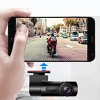 Hand holding a smartphone displaying a video of a motorcycle on a city street, with a dash cam in the foreground.